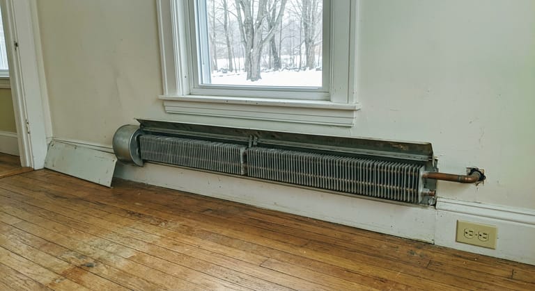 A hot water baseboard run under a window with the front cover removed, showing the finned copper element and the open damper flap.